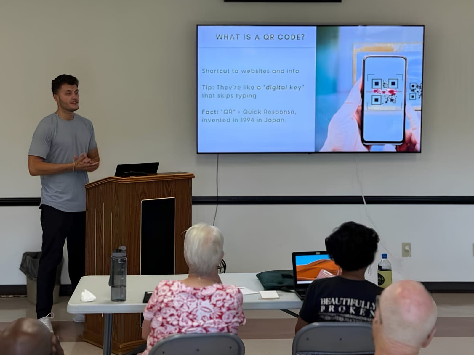 A GOLD presenter explaining what a QR code is, with the slide showing a phone scanning one
